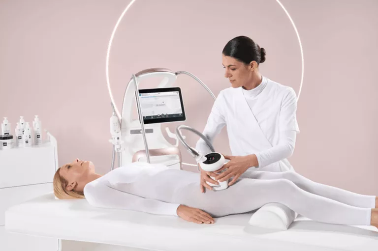 Zabieg endermologii wykonywany w gabinecie kosmetologicznym. Kosmetolog obsługuje nowoczesne urządzenie do masażu podciśnieniowego, a klientka ubrana w biały kombinezon leży na łóżku zabiegowym.