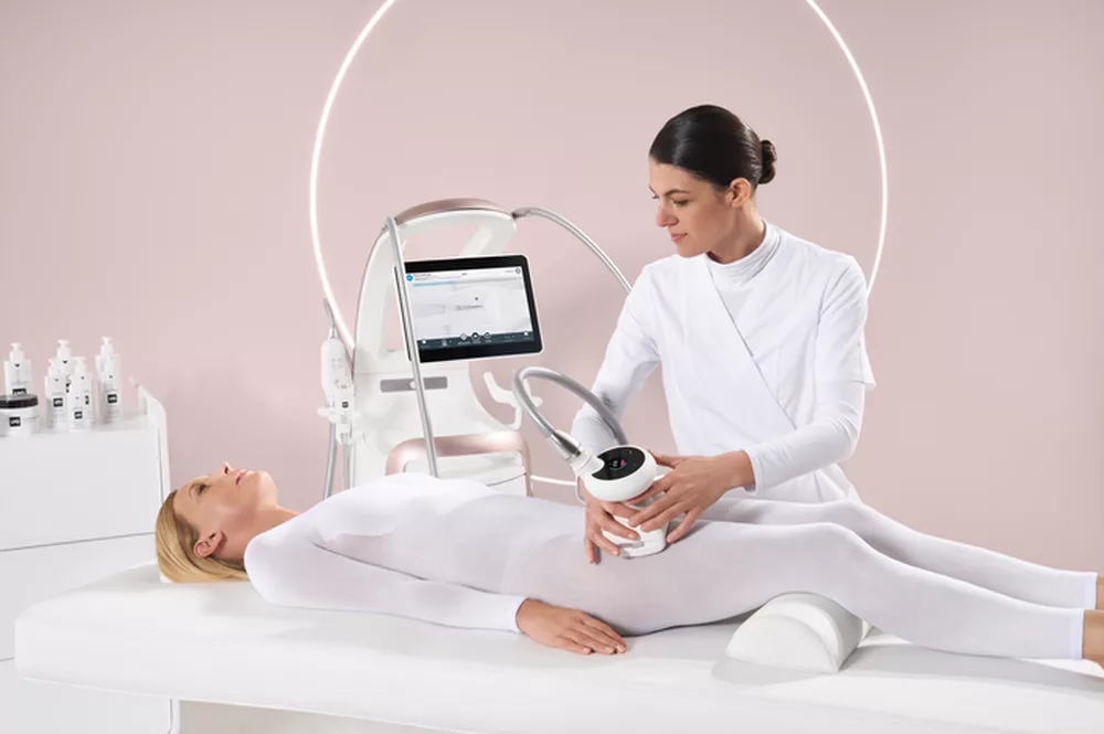 Zabieg endermologii wykonywany w gabinecie kosmetologicznym. Kosmetolog obsługuje nowoczesne urządzenie do masażu podciśnieniowego, a klientka ubrana w biały kombinezon leży na łóżku zabiegowym.