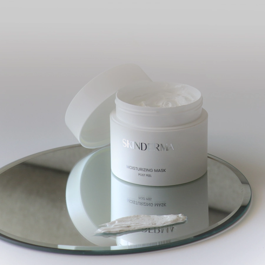 Otwarty słoik nawilżającej maski SKINDERMA stoi na okrągłym lustrze, z niewielką ilością kremu nałożoną obok na powierzchni lustra.