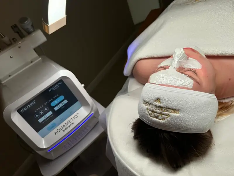 Osoba leży na łóżku zabiegowym w gabinecie kosmetycznym, z nałożoną białą maską na twarzy, podczas gdy obok znajduje się urządzenie Aquametiq Hydra Prime używane do oczyszczania wodorowego.